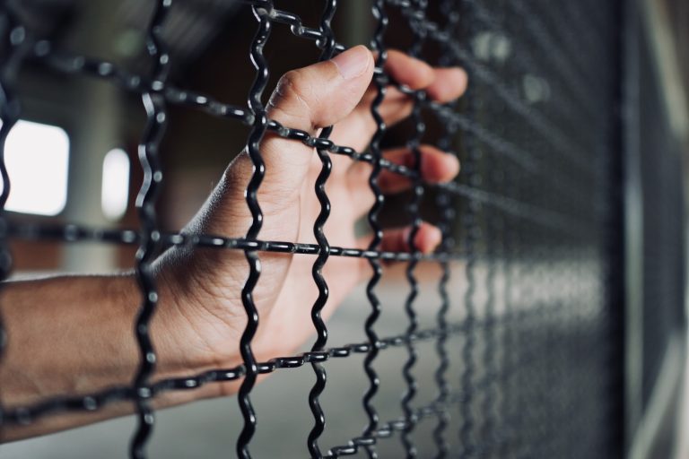Hand,Of,Prisoner,Holding,Metal,Fence,,Criminal,Locked,In,Jail