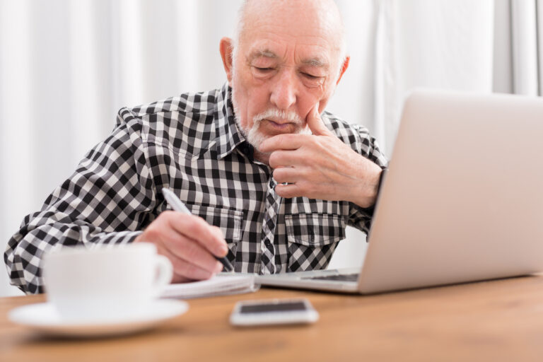 Mature,Man,Using,Laptop,And,Writing,In,Notepad,At,Home