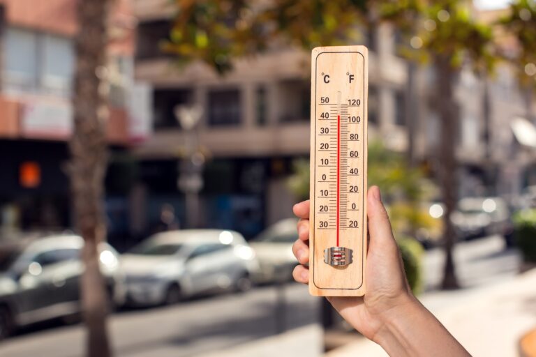 Hot,Weather.,Thermometer,In,Hand,In,Front,Of,An,Urban