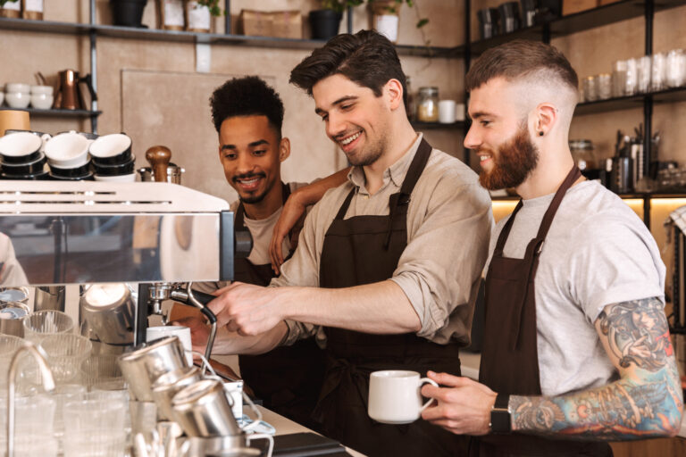 Three,Cheerful,Male,Baristas,Standing,At,The,Coffee,Shop,Counter