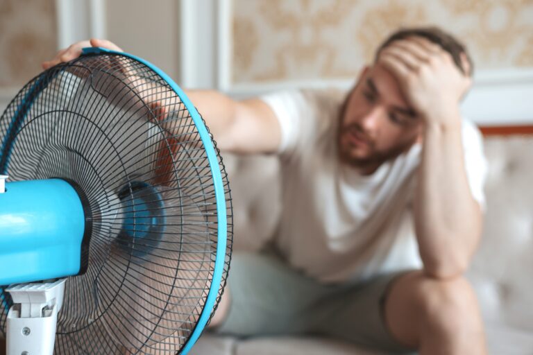 Young,Bearded,Man,Using,Electric,Fan,At,Home,,Sitting,On
