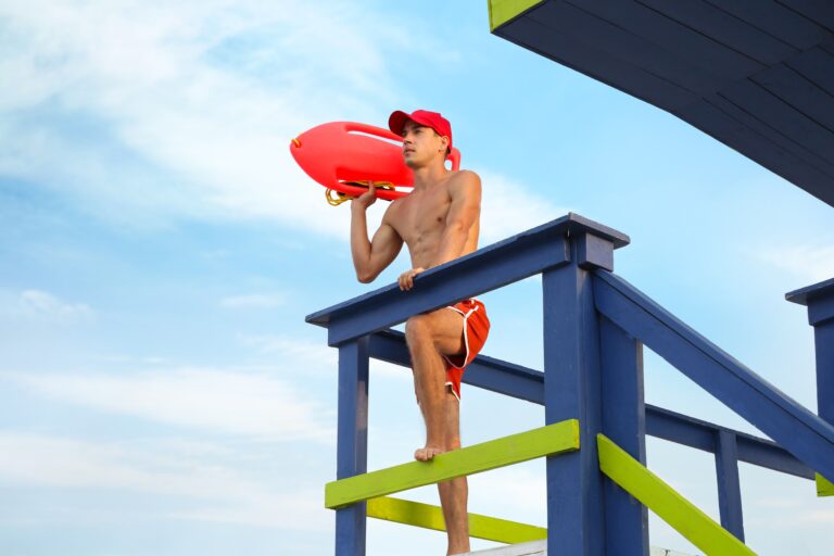 Handsome,Lifeguard,With,Life,Buoy,On,Watch,Tower,Against,Sky
