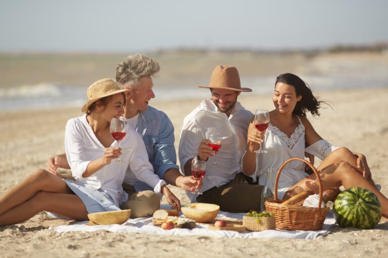 Two,Couples,Enjoying,A,Picnic,On,The,Beach,,Toasting,With
