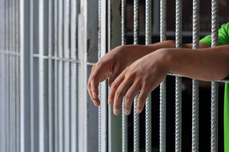 Hands,Of,A,Prisoner,Behind,Prison,Bars,On,Black,Background